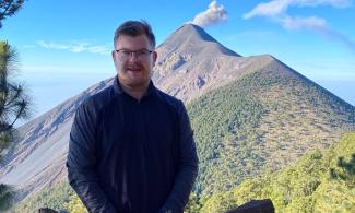 Earth Scientist Julian Pahl stands front of a volcano and blue sky. The volcano is smoking slightly.