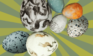 Various sizes of paper eggs are laid out, some painted blue with speckles and others abstractly decorated with black paint. Set on a light and dark green striped background.