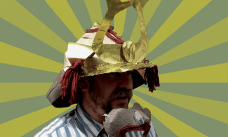 A man wears a gold and red samurai helmet, holding a matching mouthpiece with a large red mouth design. He is dressed in a blue and white striped shirt, standing against a light and dark green striped background.