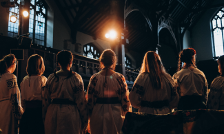 A group of women in Ukrainian clothing stand in a line with their backs to the camera as lights shine down upon them.