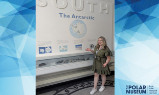 Member of staff in a green dress standing in front of a case labelled 'South: The Antarctic' which contains fossils and rocks. 