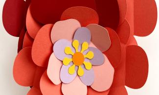 A hand holds a bouquet of paper flowers with deep red outer petals and pink and purple inner petals. The centres are orange and yellow, with wooden sticks as stalks.