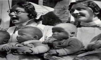 Two nurses dressed in traditional white uniforms and caps smile as they each hold two babies on their laps, making a total of four infants. The babies are dressed warmly in knitted clothing and are sitting side by side, looking in various directions. The photo appears to be black and white.