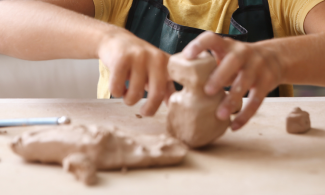 A child wearing a yellow t-shirt and an apron sits at a table and shapes some clay.