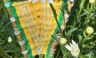 A piece of yellow and green weaving between two bits of woods, propped up in a garden next to a daisy.