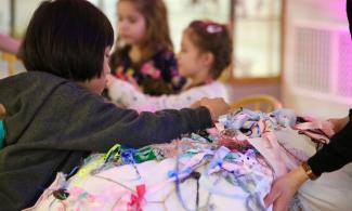 alt="a child with dark hair and a grey jumper reaches across a patchwork quilt to tie a ribbon onto it"