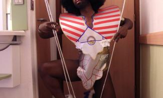 Harold Offeh, perched on one knee, wearing a short-sleeved silver and red costume, and bright blue high heels. He also wears a thick, curly black wig.