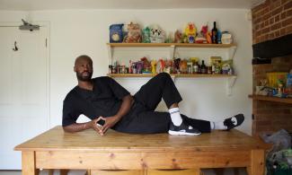 Harold Offeh lounging on a kitchen table, propped on one elbow, and looking at the camera.