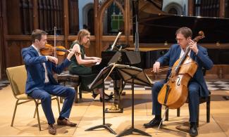 Three people sit on chairs in a church, playing string instruments.