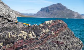 A coastal scene where a large rock is in the close-up foreground with red and white mineral patches within it. A geological hammer rests on top of the rock. Behind the rock is blue sea and sky, with a high craggy, rocky hill on the other side of the sea. Beyond this are mountains in the far distance.