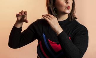 A woman wearing a black dress, with her hands raised, as if pretending to play a flute.
