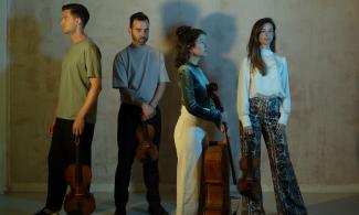 Four people standing in a room, holding string instruments, and looking in various directions.