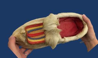   A view from above of a shoe, made from off-white, fluffy reindeer fur, held by two hands. The shoe is bright red inside, and has stripes of red, yellow and blue felt on the front, and a curled up toe.