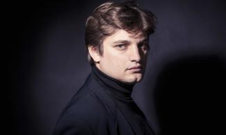 A man with thick brow hair looking at the camera. He is dressed all in black.