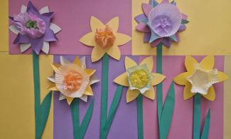 A colorful arrangement of paper daffodils crafted from cupcake liners and pastel paper petals on a background of pink, purple, and yellow paper. Each daffodil has a unique center in colors like orange, pink, yellow, and purple. Long green paper stems complete the floral display.