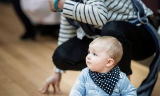 alt= "a baby in a blue shirt and neckerchief sits on the ground in front of his mum who is crouching behind him"