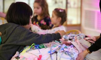 alt= "a child with black hair reaches over a pile of colourful ribbons and string"