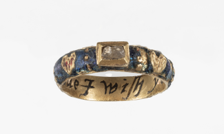 A gold and enamel ring decorated on either side of a rectangular crystal with hearts. Inside the band is an inscription.