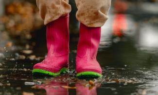 A person wearing vibrant pink rain boots with green soles stands in a puddle on wet pavement. Fallen leaves and raindrops are scattered around.