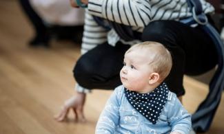 alt='a baby sat on the floor in a blue shirt, his mother crouches behind"
