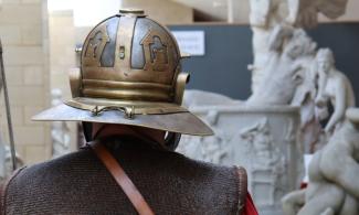 A Roman soldier with his back turned to us walks through a gallery of sculpture.
