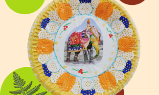 A paper plate has been painted in gold paint to form a background colour, on top is a white doily which has some elements coloured in orange and blue around the rim. In the centre is a photograph image of a brightly decorated Indian elephant. 