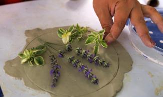 A hand arranges sprigs of lavender and small green leaves on rolled-out clay.
