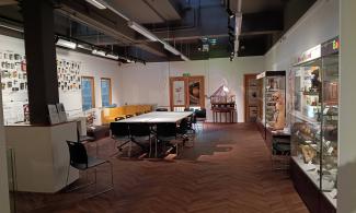 alt="the image shows the Learning Gallery at the Whipple Museum. It has brown wooden floor, white walls and a white table in the middle surrounded by thirteen black chairs with a yellow sofa in the back left corner. Displays showing Scientific displays, from calculators to anatomy models line the walls"