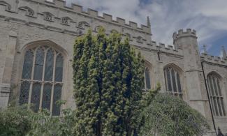 A view of the church from the side, with several windows visible and a blue sky with wispy clouds above. In the foreground is a tall slim hedge, with trees surrounding.