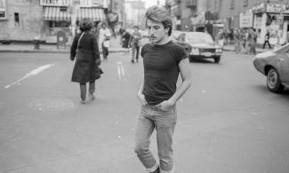 Black-and-white photograph of a young man with swept-back hair and a moustache walking along a busy city street. He wears a fitted dark T-shirt, cuffed jeans and boots, with his hands in his pockets and his eyes lowered. Cars, pedestrians and shopfronts recede behind him.