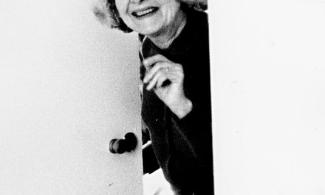 Black and white photograph of Helen Ede wearing glasses and smiling as she peeks around a partly open door.