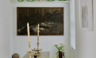The Kettle’s Yard dining room, with brass candlesticks on a wooden table, a striped textile draped over a bench, and a large painting beneath green and white plates.