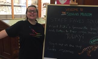 A Sedgwick Museum staff member grins and throws their arms out wide as they stand next to a sign advertising staff favourites tours. 