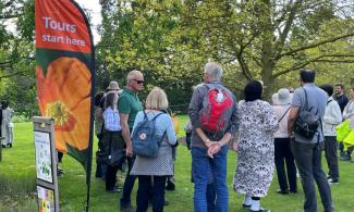 A group of people wait by an orange flag on a lawn, which has the text 'Tours start here', with trees in the background.