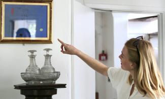 A person in a white shirt points to a blue abstract painting in a gold frame inside Kettle’s Yard. Two glass decanters sit beneath it on a dark, carved wooden pedestal.