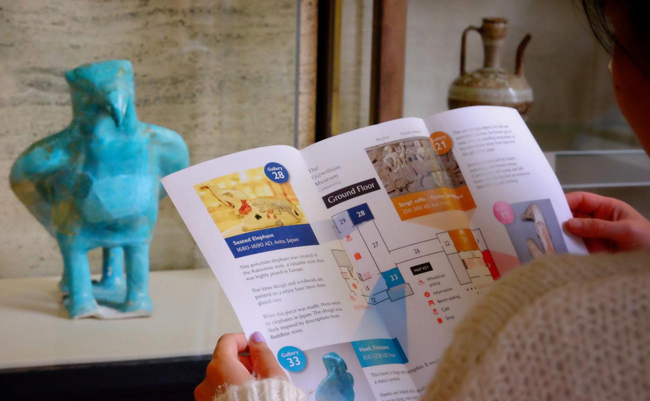 Someone holds open a Museum Walk leaflet in front of a turquoise statue of a hawk at the Fitzwilliam Museum.