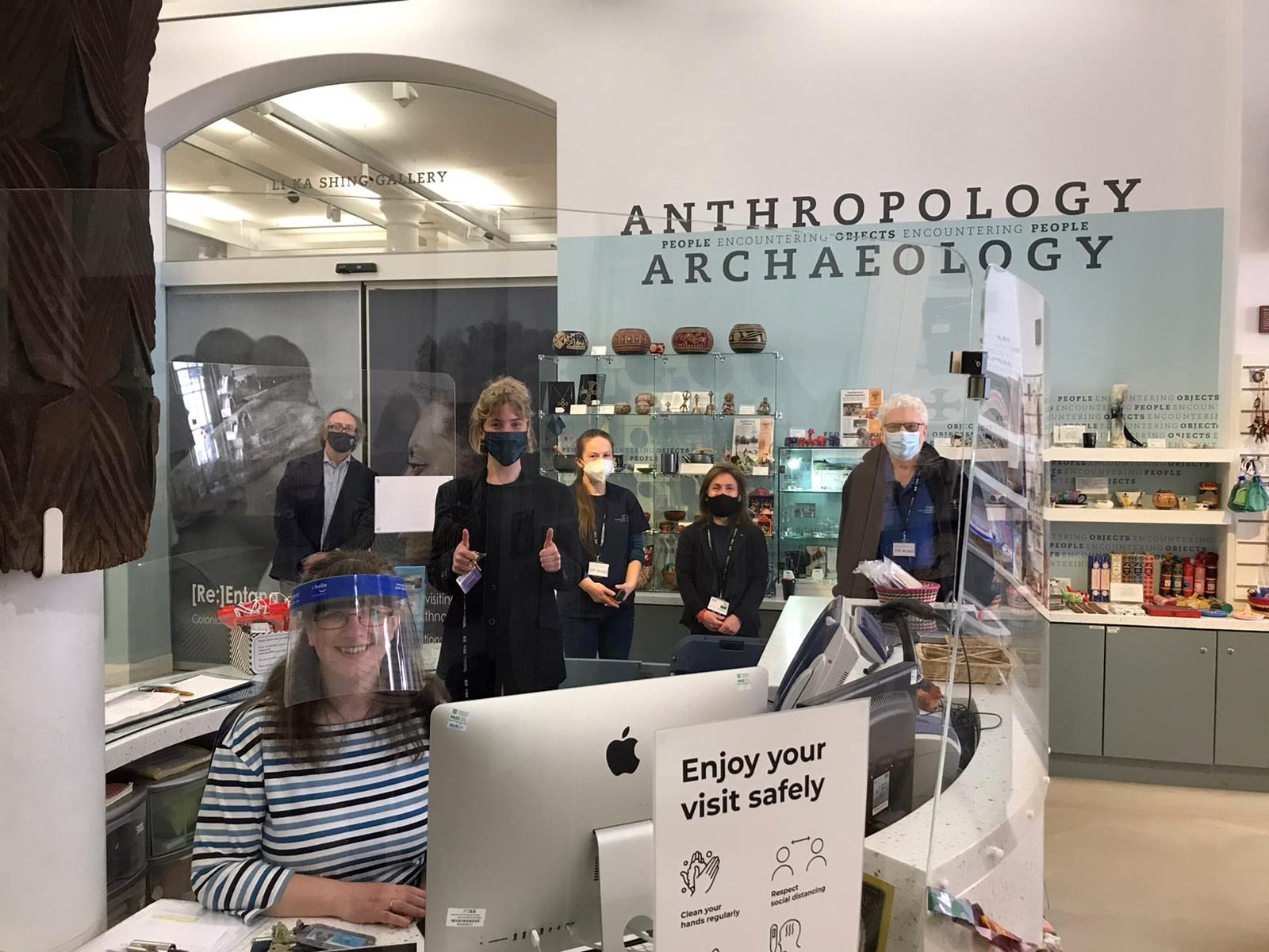 Six members of staff from the Museum of Archaeology and Anthropology, wearing face masks or visors, are socially-distanced gathered around the welcome desk. One is giving a thumbs up.