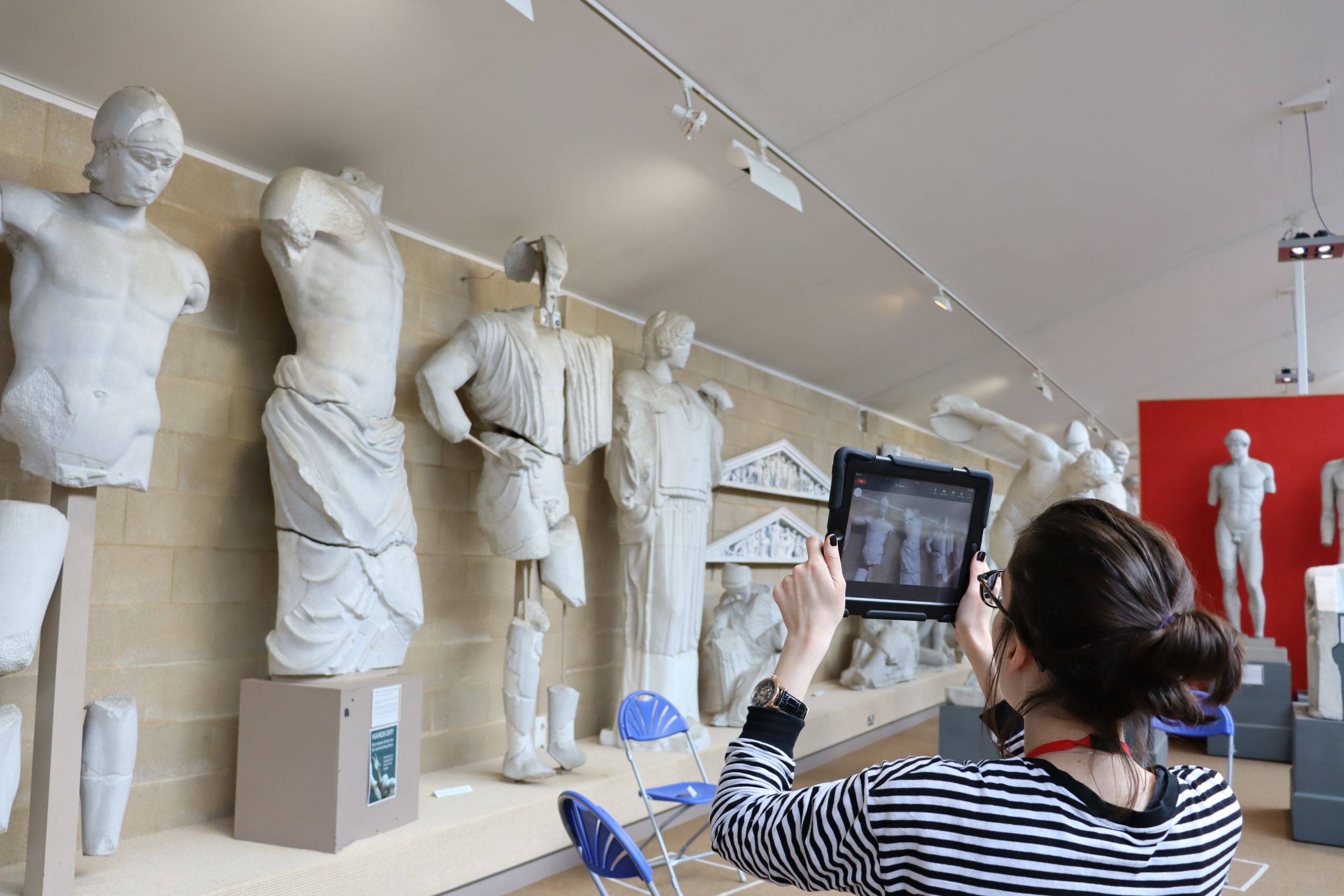 The Museum's Education & Outreach Coordinator, a woman with dark hair, holds up a tablet computer in front of a row of large white plaster statues