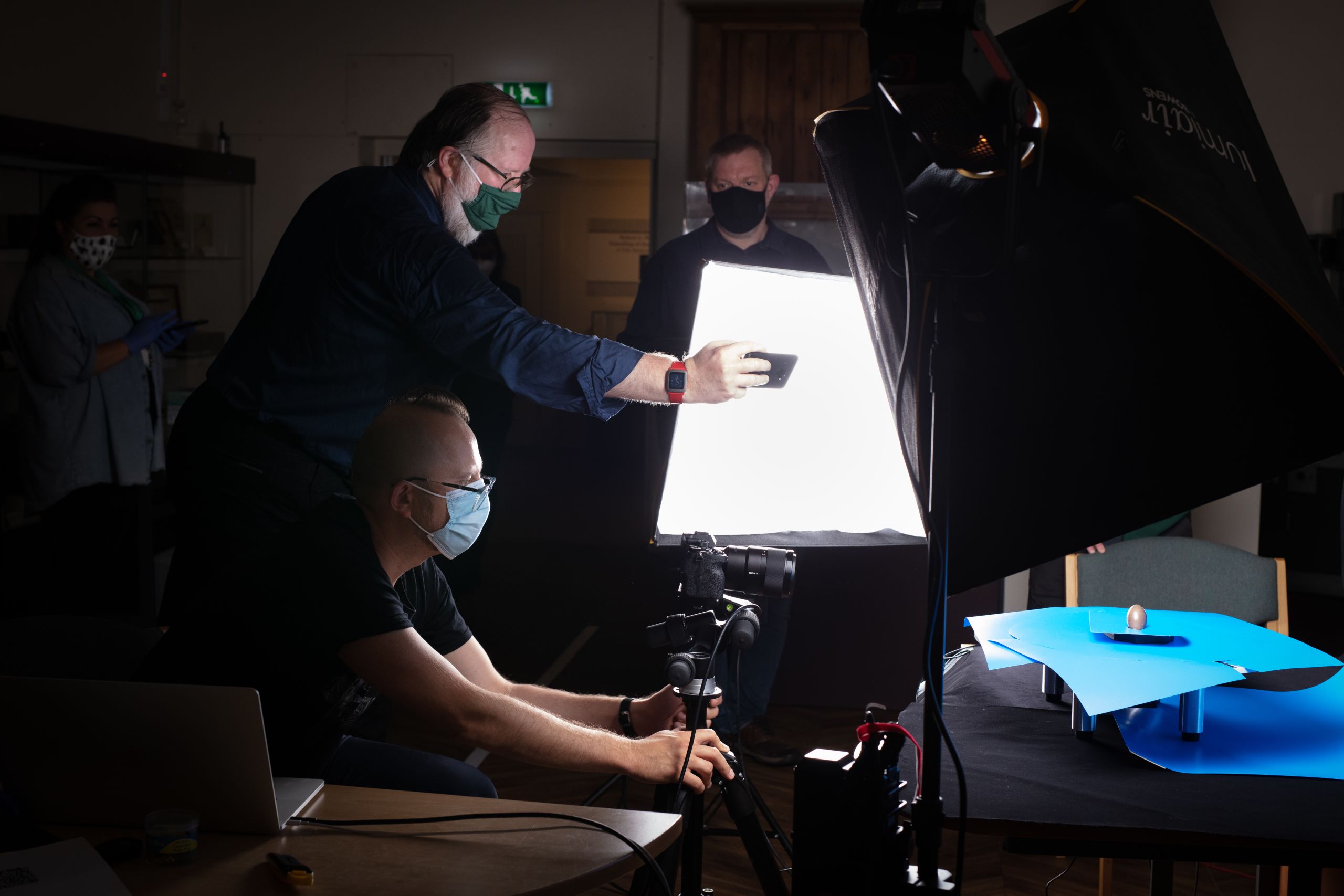 Three men wearing masks are gathered with photographic equipment around a brightly lit, tiny brown egg.