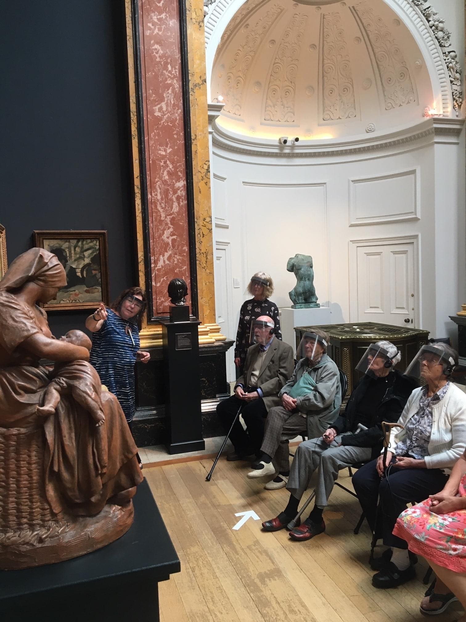 Four elderly people wearing visors, plus their support worker, sit in a museum gallery watching a museum educator as she points to a statue of a Madonna and Child
