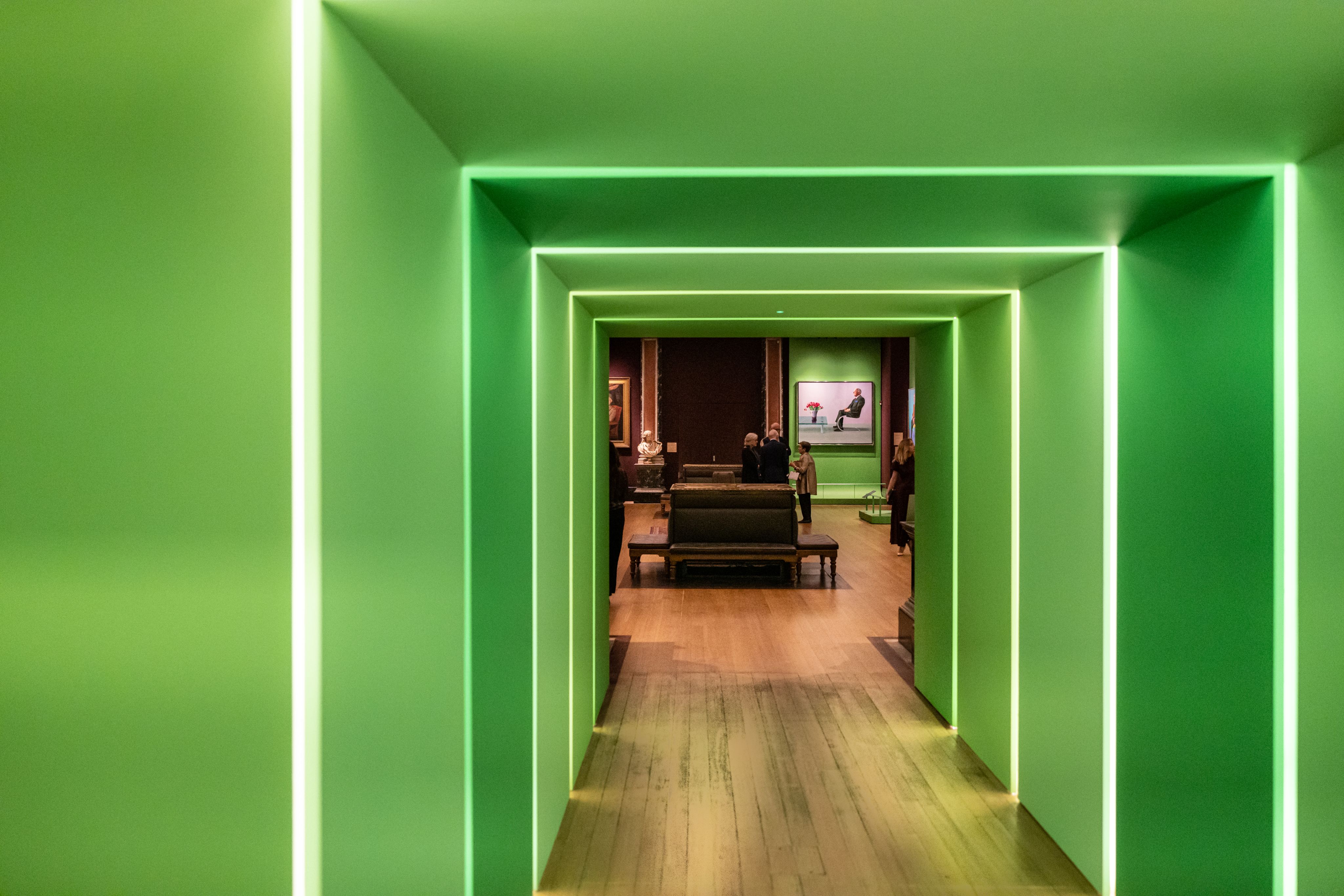 View through a bright green tunnel into the Fitzwilliam Museum's Gallery 3, where a David Hockney painting of a man sitting opposite a vase of tulips is exhibited