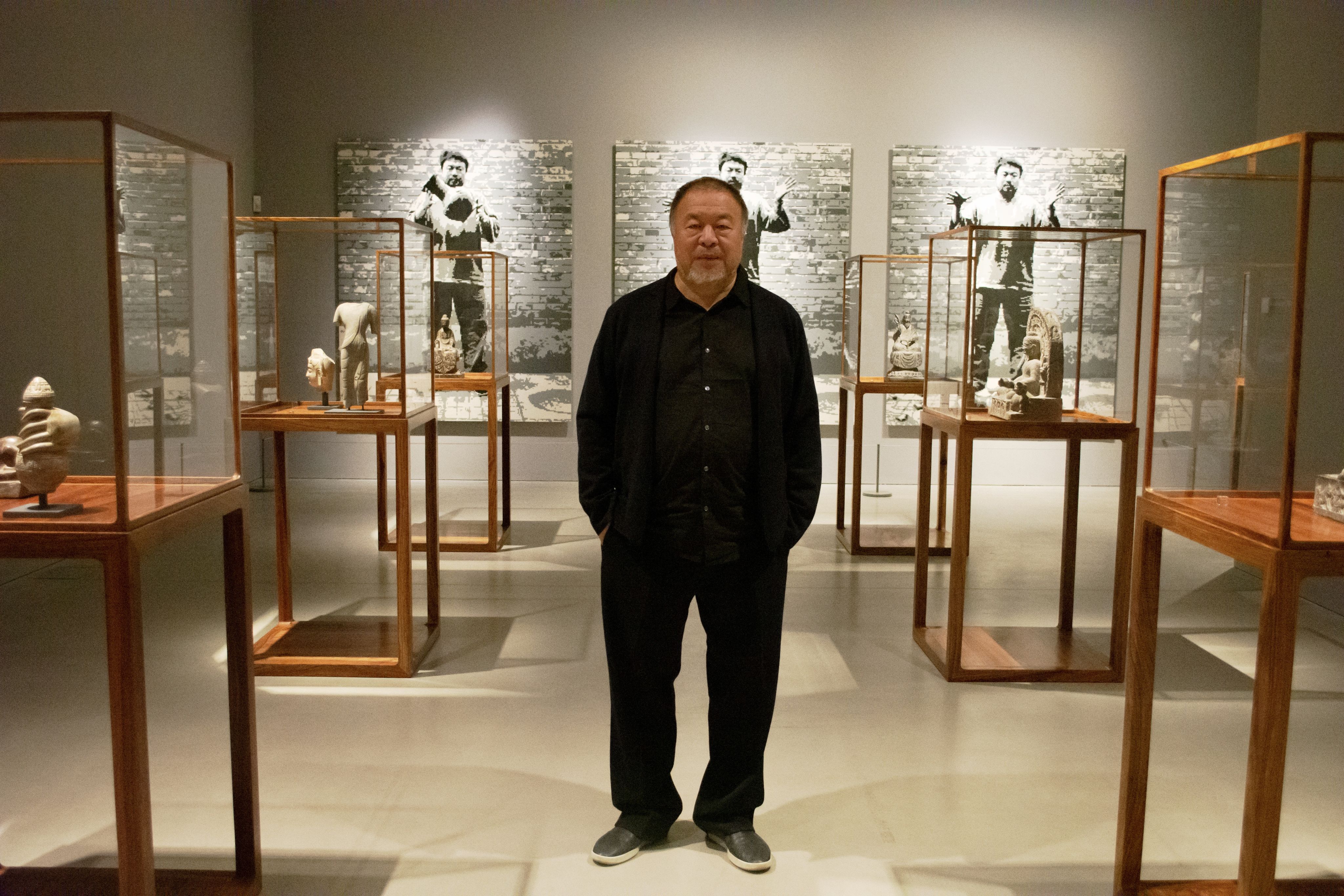 Ai Weiwei stands in the middle of a gallery at Kettle's Yard. with display cases exhibiting sculptures and prints along the back wall