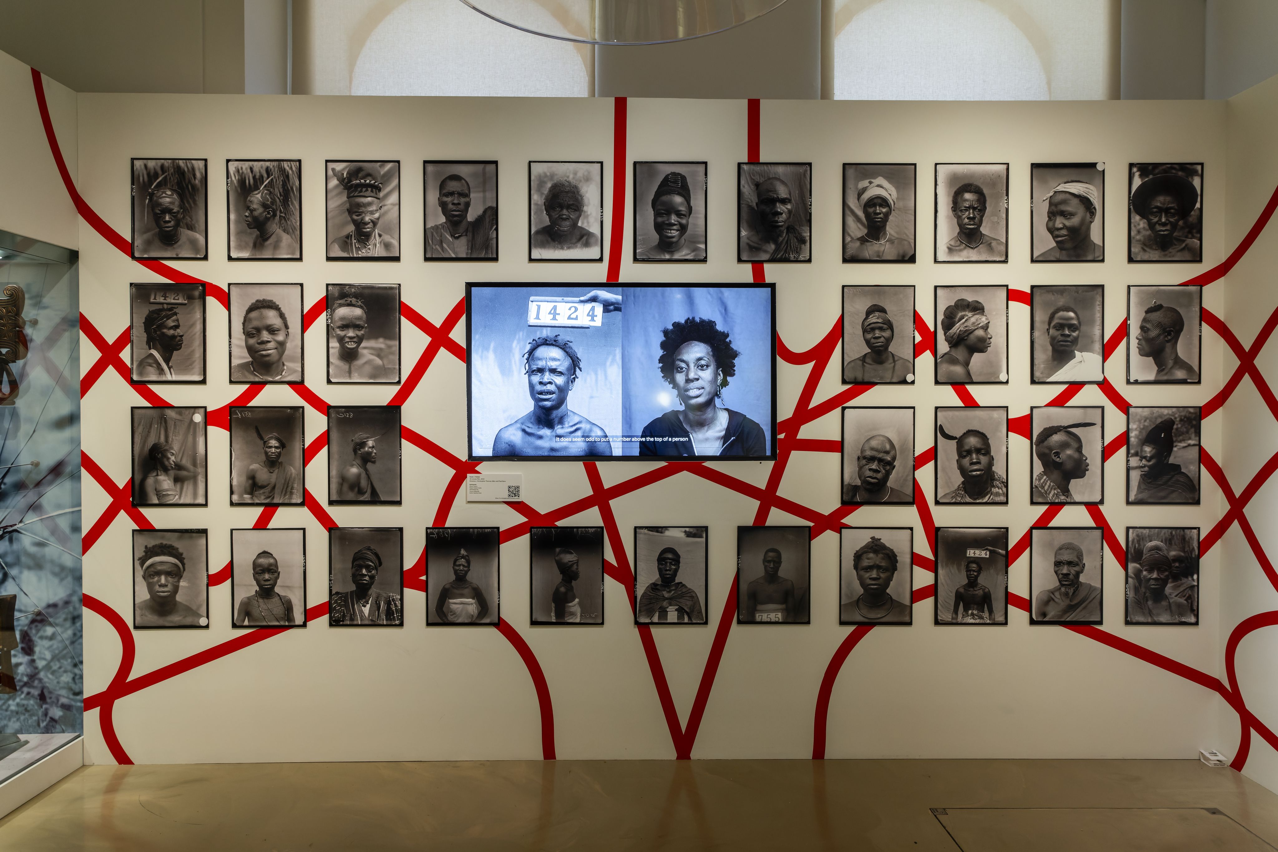 View of a gallery wall. Historic black-and-white photographs are linked by a red design. In the centre a video screen plays a film.