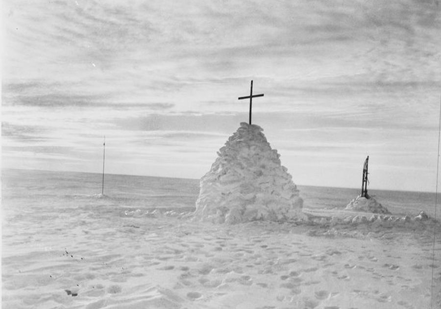 A snow cairn with a cross on top in the foreground, a smaller snow mound with sledge standing upright to one side behind. A pole stands in the snow to the other side.