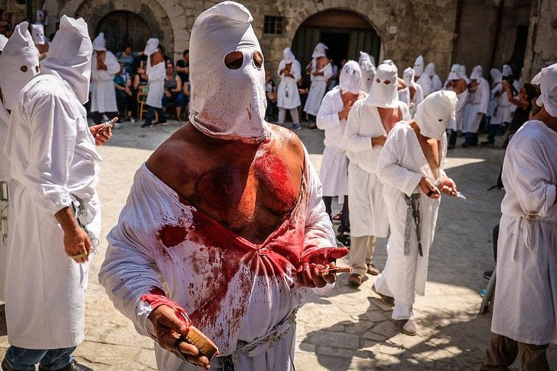 A hooded man stands with his short torn and chest bare, with blood all over his clothes and body. Behind him are a group of more hooded men.