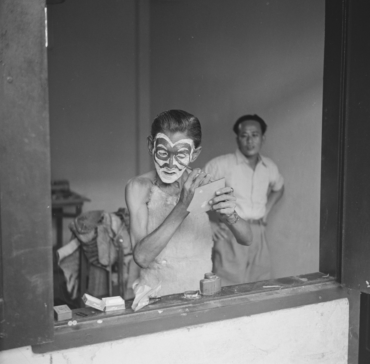 The spirit-medium applies make-up or greasepaint. An older man wearing a cloth apron, he is standing at an open window with a second man behind him.
