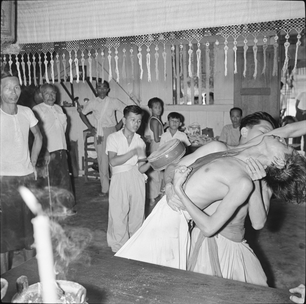 Inside the temple, the spirit-medium (a young man wearing a white apron and silk stomacher, falls back into the arms of a second man, similarly dressed. Behind them is a boy with a drum and a small group of men, boys and a woman. There is a candle in the foreground and a fringed cloth hangs from the ceiling.