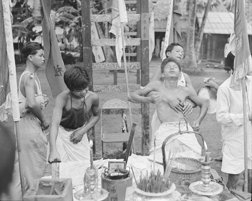 A group of young spirit mediums in white trousers are at the foot of a sword bridge, of which only the lower ladder is visible. On the right, one medium has fainted in the arms of another. In the foreground is a table with incense sticks, candles and a basket. On the left, another medium in a dark apron leans forward to reach for a sword.