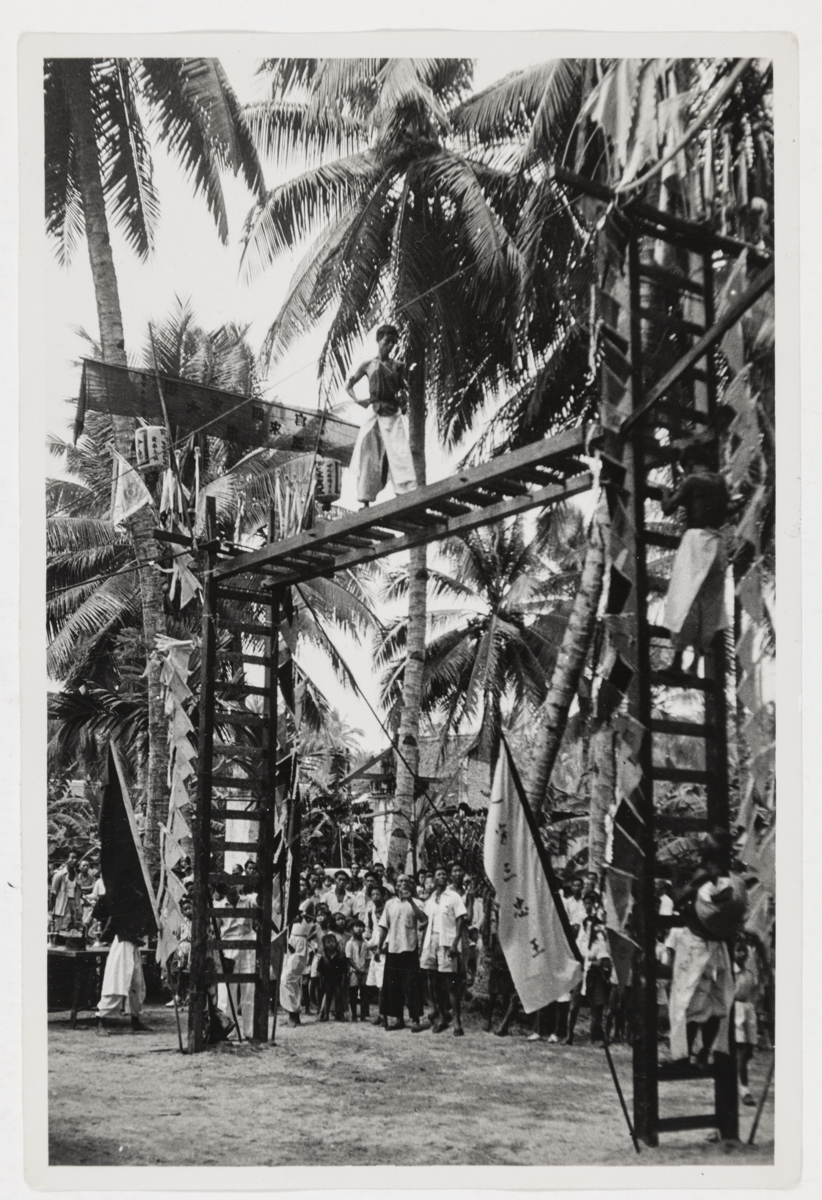 A medium in white trousers and vests stands at the top of an open-air sword bridge, while two more men climb the right-hand ladder. Onlookers watch from the ground. 