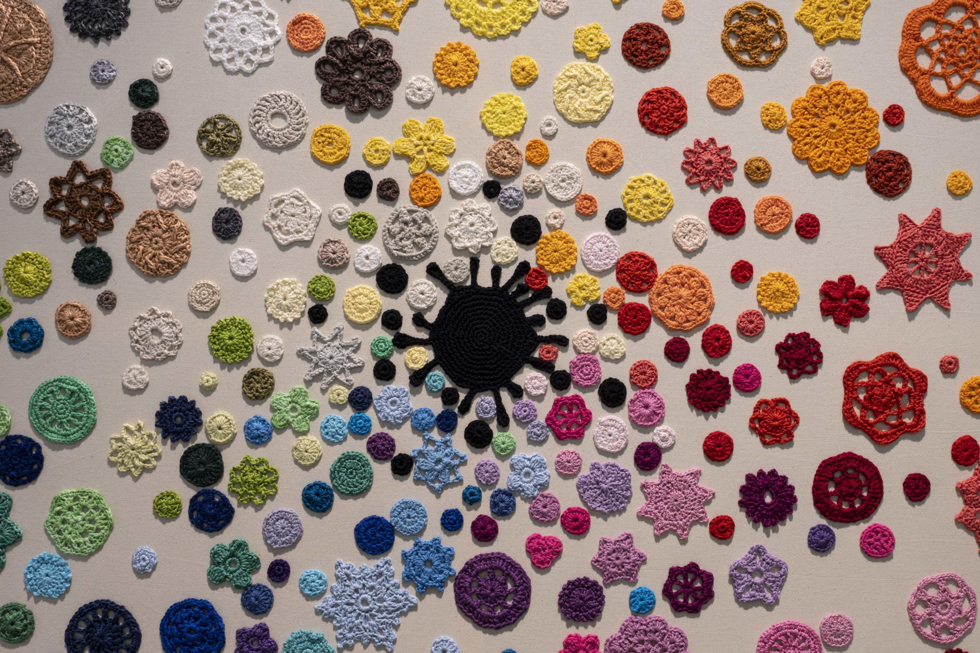 Close up shot of the 'Kaleidoscope' installation, showing dozens of multicoloured crocheted flowers arranged in a rough colour wheel around a black central blob. Mounted on calico.